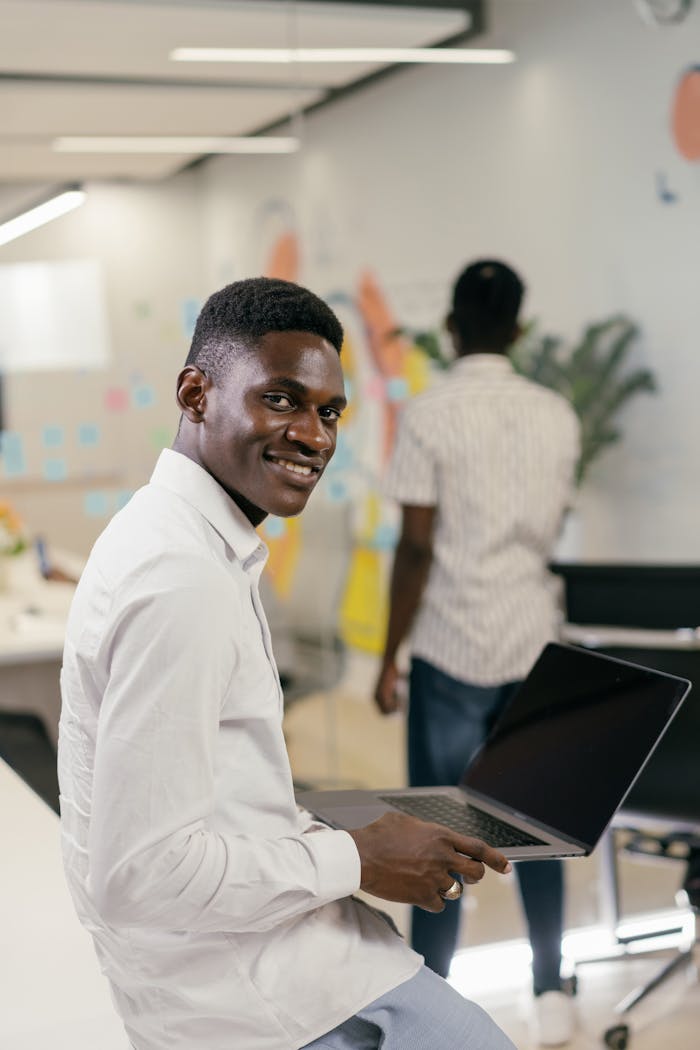 A young man in a white shirt smiling while holding a laptop in an office setting.
